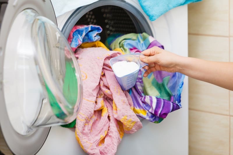 Uma pessoa colocando sabão em pó em uma máquina de lavar roupas. A máquina tem porta vertical e já está com roupas, de cama, dentro. 