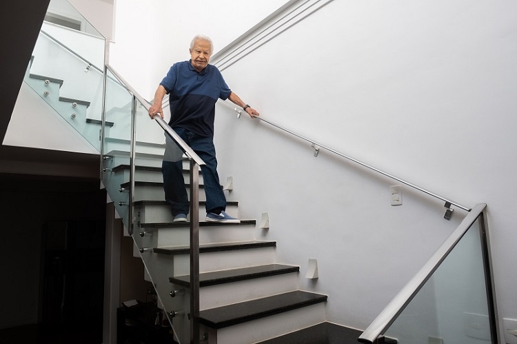 Cid Moreira desce as escadas de seu apartamento, apoiado nas barras de proteção no corrimão.