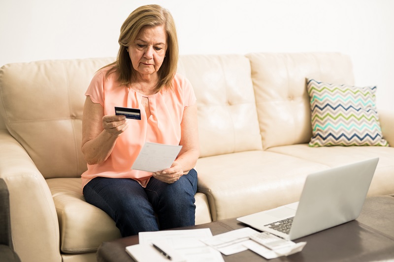 Uma mulher com mais de 50 anos olha seu cartão de crédito e pensa nas contas que não pode pagar. Imagem ilustra notícia sobre salário mínimo atualizado.