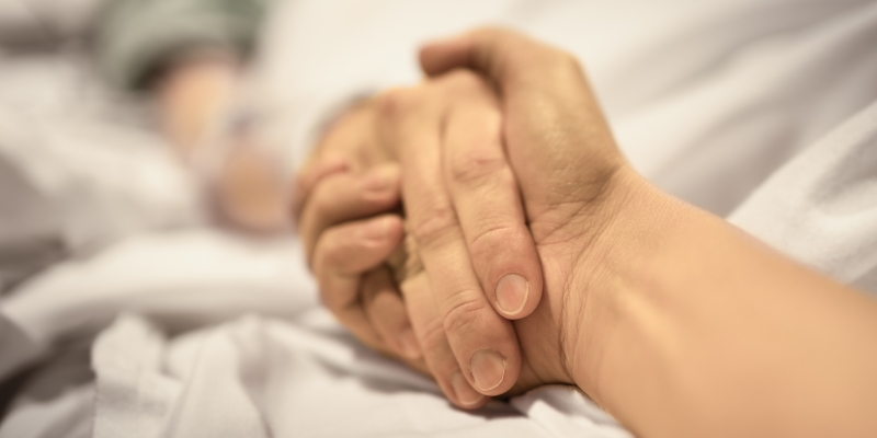 Duas mãos se segurando sobre um leito de cama ou cama hospitalar. Imagem ilustrativa para o conteúdo sobre falar sobre morte