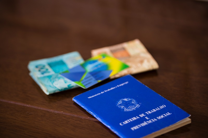 Uma carteira de trabalho fechada sobre uma mesa. Ao seu lado, um cartão do benefício do INSS, com notas de 100 e 50 reais. Imagem para ilustrar a matéria sobre perder o emprego.