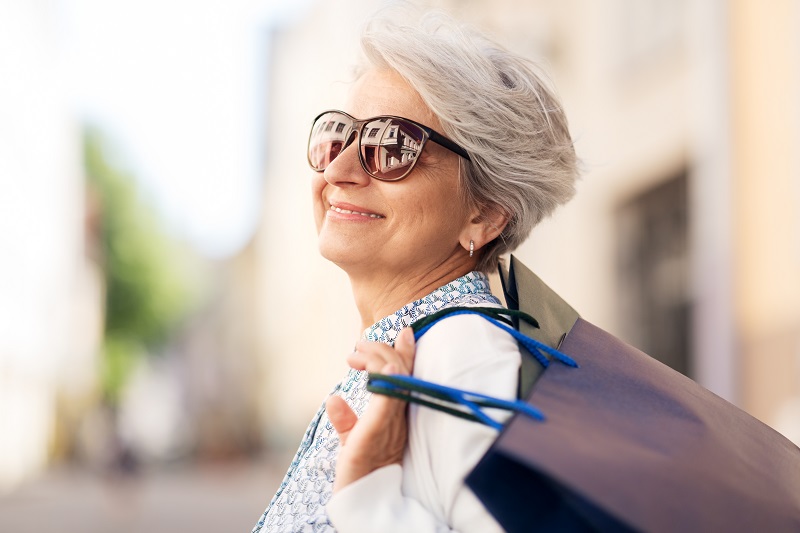 Uma mulher com mais de 60 anos sorri com sua sacola de compras. Imagem ilustra a matéria sobre como economizar roupas na primavera.