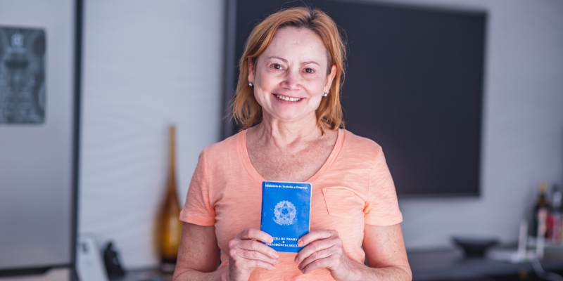 Uma senhora de cabelos claros, sorrindo, em pé e segurando uma carteira de trabalho. Imagem para ilustrar a matéria sobre Reforma da Previdência.
