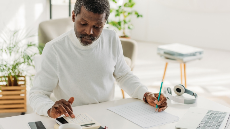 Um homem sentando em uma mesa, mexendo em uma calculadora e anotando informações sobre o seu planejamento financeiro.