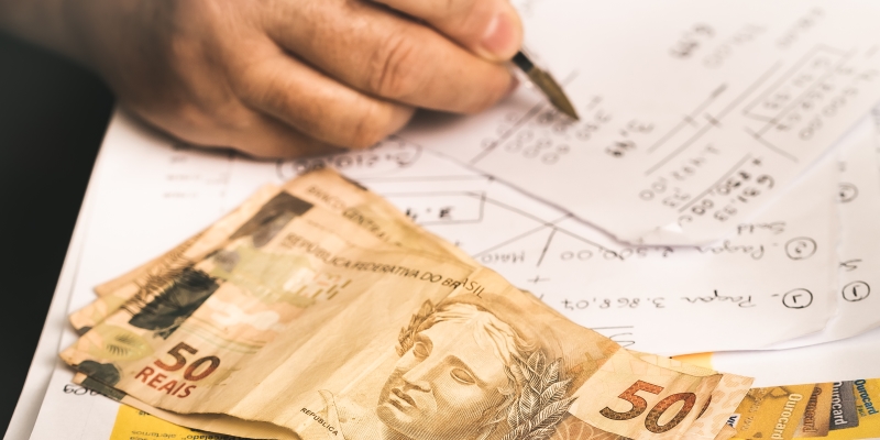 Uma pessoa fazendo contas com caneta e papel e com notas de 50 reais sobre a mesa. Imagem para ilustrar a matéria sobre renegociação de dívidas para MEIs.