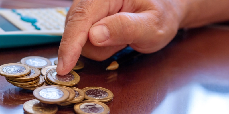 Uma mão masculina mexendo em moedas de um real, sobre uma mesa. Ao lado, uma calculadora e um lápis. Imagem para ilustrar a matéria sobre dinheiro esquecido.