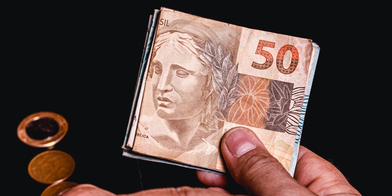 Uma mão masculina segurando notas de dinheiro. Ao fundo, moedas. Imagem para ilustrar a matéria sobre linha de crédito para microempresas.