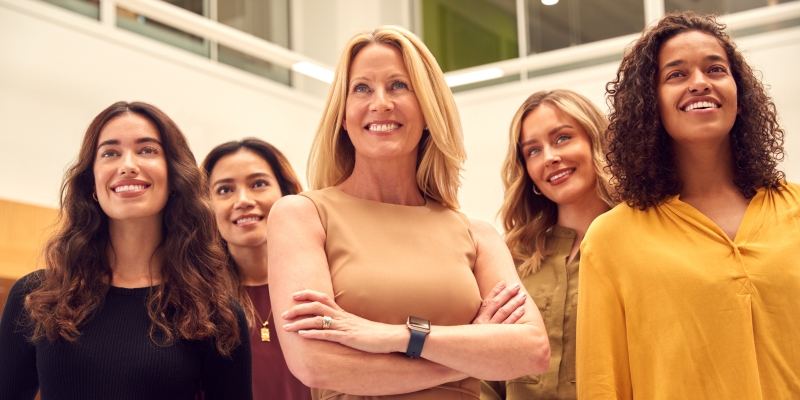 Cinco mulheres, sendo uma centralizada e com cerca de 50 anos, em um escritório, em pé e olhando para frente. Imagem para ilustrar a matéria sobre preconceito contra a mulher no mercado de trabalho.