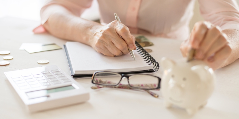 Uma mulher idosa fazendo anotações em um caderno e colocando uma moeda e um cofrinho. Imagem para ilustrar a matéria sobre como ganhar mais dinheiro.
