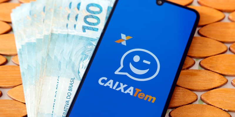 Notas de 100 reais e um celular sobreposto, com o aplicativo da Caixa Tem aberta. Imagem para ilustrar a matéria sobre o microcrédito SIM Digital.