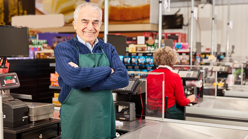 Homem 50+ posa olhando para a foto, com um avental de trabalho em um caixa de mercado. A imagem ilustra o artigo sobre longevos.