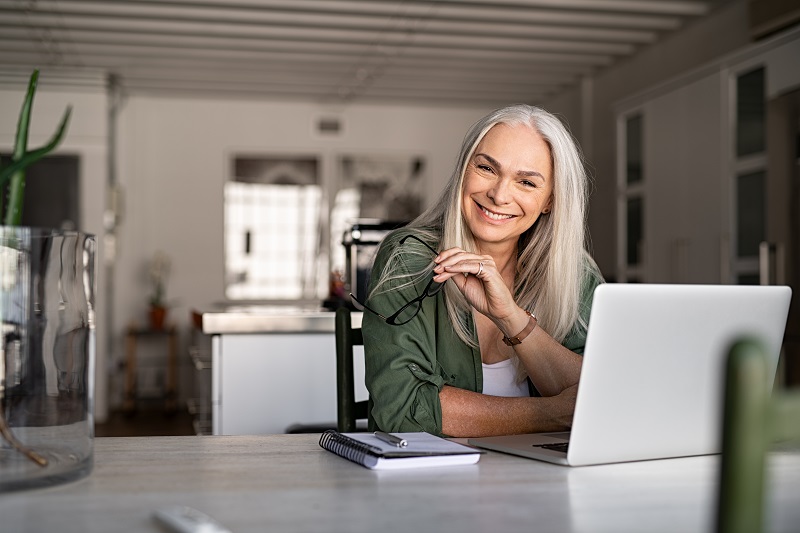 Mulher 50+ sentada à frente de uma mesa e com um computador aberto. A imagem ilustra o artigo sobre longevos.