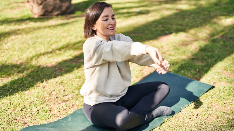 Uma mulher de meia idade sentada de pernas cruzadas e sobre um tapete de yoga em uma parque gramado. Ela está alongando o braço e sorrindo. Imagem para ilustrar a matéria sobre 10 hábitos.