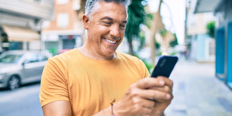 Um homem feliz olhando para o celular e sorrindo. Imagem para ilustrar a matéria sobre como apostar pelo celular.