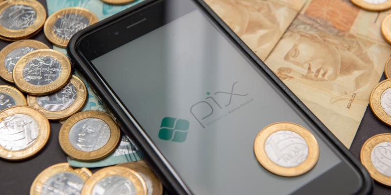 Um celular com sobre uma mesa. Nele, a imagem, com a logo do PIX. Ao redor do celular, dinheiro e moedas de R$ 1. Imagem para ilustrar a matéria sobre o novo golpe do PIX.