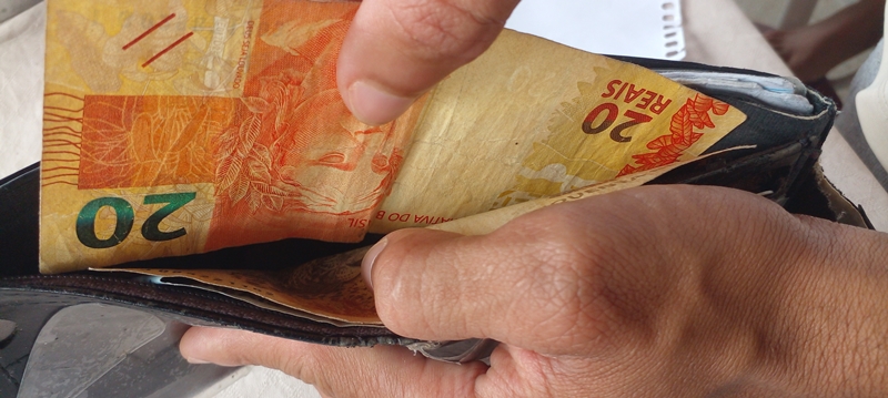 Uma mão masculina abrindo uma carteira. Dentro dela, há notas de dinheiro e ele puxa da de 20 reais. Imagem para ilustrar a matéria sobre pesquisa Datafolha sobre problemas financeiros.