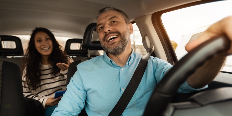 Um motorista de aplicativo conversando com uma passageira, no banco de trás, enquanto os dois estão no carro. 