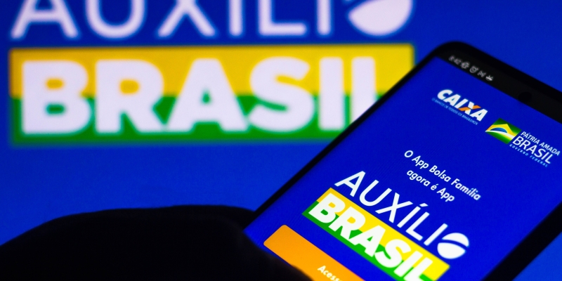 Um celular com o aplicativo do Auxílio Brasil aberto. Ao fundo, uma tela de computador com a logo do Auxílio Brasil. Imagem para ilustrar a matéria sobre novos cartões do Auxílio Brasil