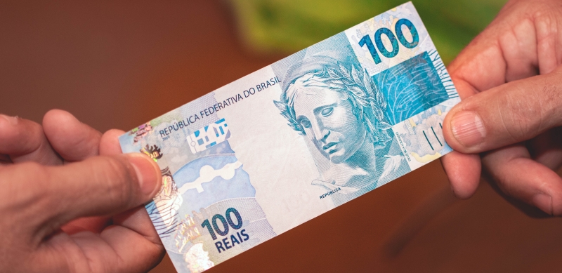 Close de mãos. Uma pessoa entregando uma nota de 100 reais para outra. Imagem para ilustrar a matéria sobre Dinheiro emprestado.