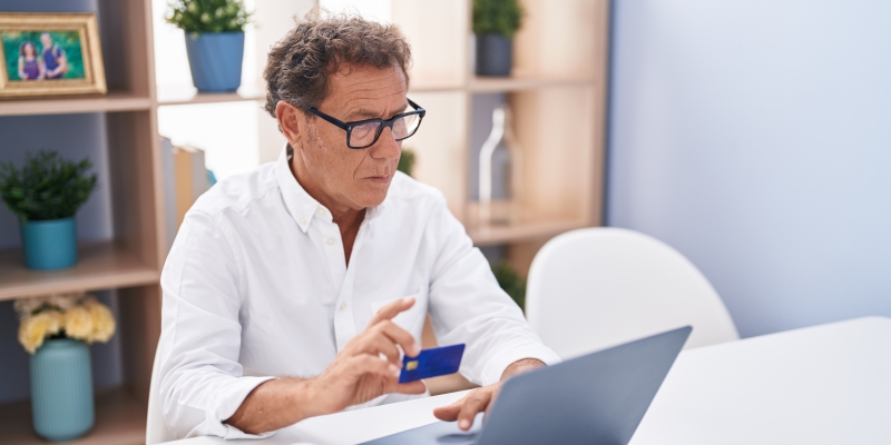 Um homem fazendo compras na internet, em frente a um computador portátil, segurando um cartão de crédito. Imagem para ilustrar a matéria sobre consumismo.