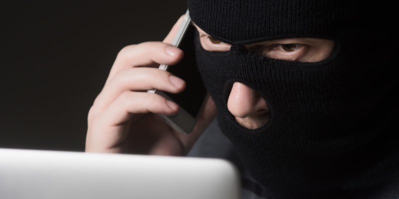 Um criminoso, ligando para uma pessoa e olhando para um computador. O homem usa máscara no rosto e está aplicando o golpe do saque-aniversário.