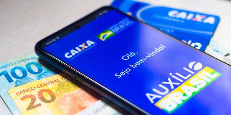 O logotipo do Auxílio Brasil exibido em um smartphone, ao lado de notas de dinheiro reais brasileiras. Imagem para ilustrar a matéria sobre o consignado do Auxílio Brasil.