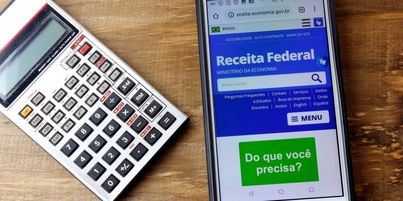Logo da Receita Federal do Brasil na tela do Smartphone. Celular está sobre uma mesa, ao lado de uma calculadora. Imagem para ilustrar a matéria sobre lote residual do IRPF.