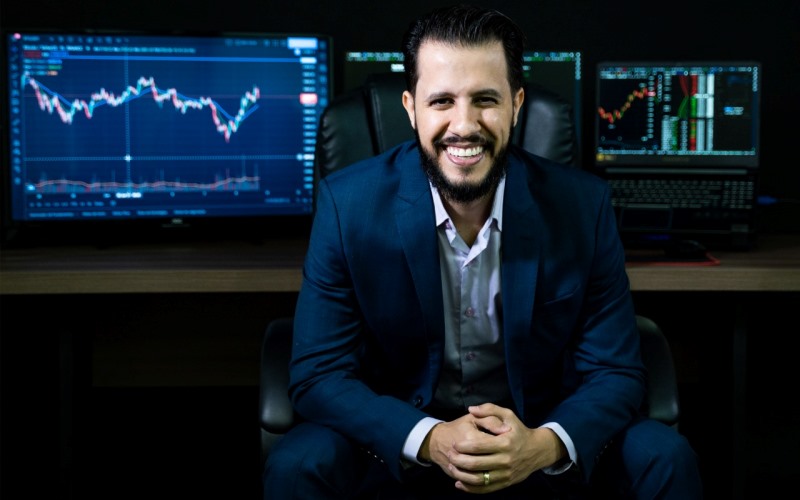 O educador e especialista financeiro Danilo Ferraz sentado em uma cadeira. Ao fundo, imagens de gráficos de finanças. Imagem para ilustrar a matéria sobre preguiça e finanças.