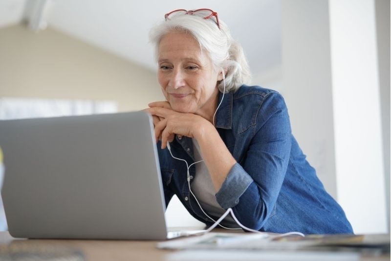 Uma mulher idosa de cabelos brancos sorrindo enquanto olha para o computador. Ela está usando fones de ouvidos. Imagem para ilustrar a matéria sobre mentiras sobre longevidade.