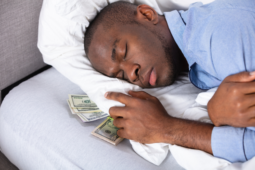 Imagem de um homem dormindo em uma calma com dinheiro debaixo do seu travesseiro. Imagem para ilustrar a matéria sobre poupar certo.